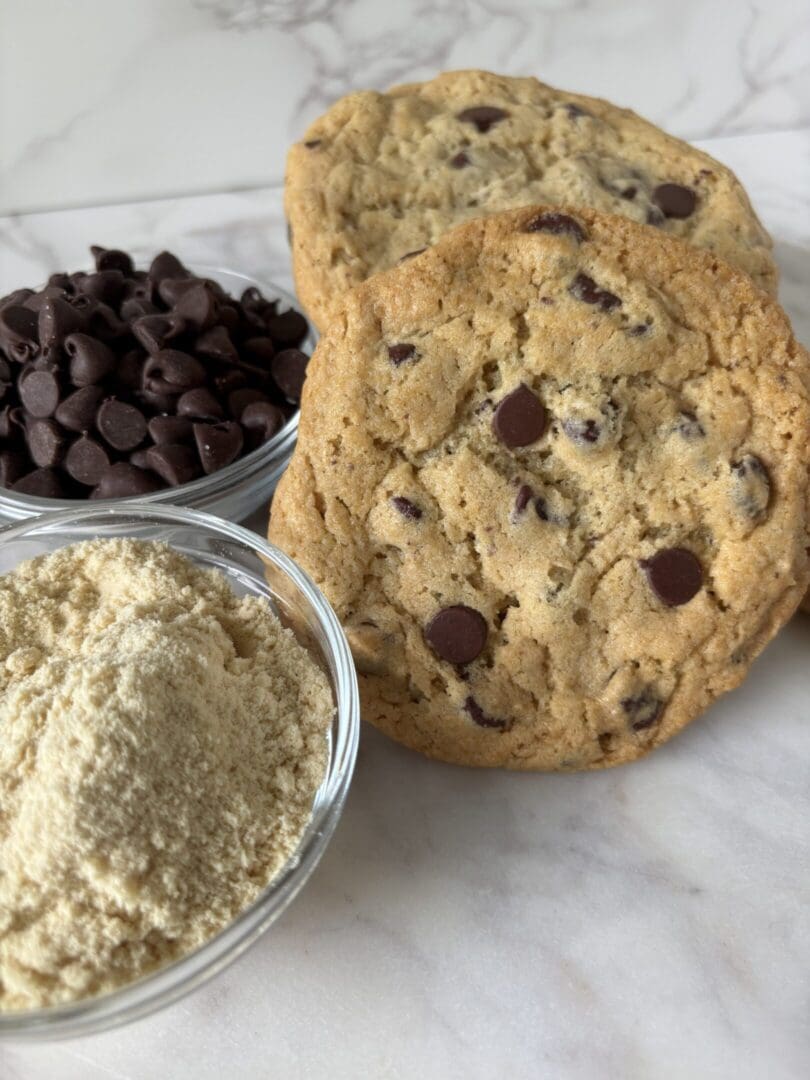 Two chocolate chip cookies with bowls of flour and chocolate chips.