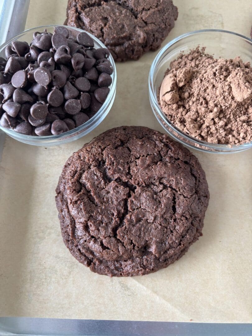A rich chocolate cookie with bowls of chocolate chips and cocoa powder.