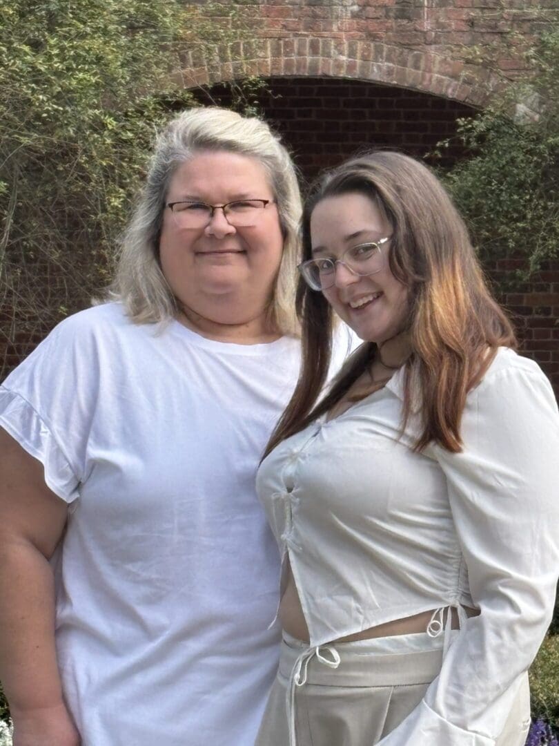 Two smiling women standing outdoors, dressed in white tops.